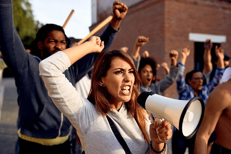 A group of angry protesters raising fists, with a woman shouting into a megaphone in the foreground during a political demonstration.