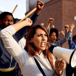 A group of angry protesters raising fists, with a woman shouting into a megaphone in the foreground during a political demonstration.
